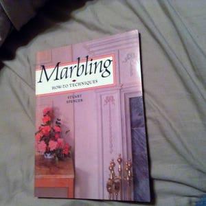 May include: A book titled "Marbling: How-To Techniques" by Stuart Spencer. The cover features a pink and white floral arrangement, a fireplace with gold accents, and a light pink background.