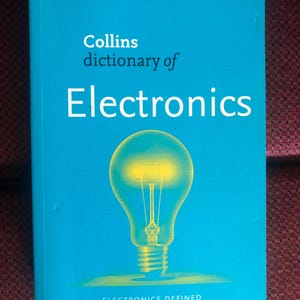 May include: A turquoise book titled "Collins dictionary of Electronics" with a yellow lightbulb graphic. The book cover also includes the text "Electronics Defined and Explained". The Collins logo is in the upper left corner.