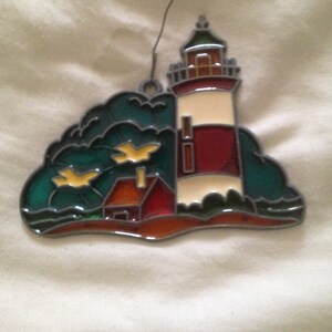 May include: A stained glass ornament depicting a red and white lighthouse with a small house in front of it. The lighthouse is on a small island with green trees and yellow birds flying overhead.