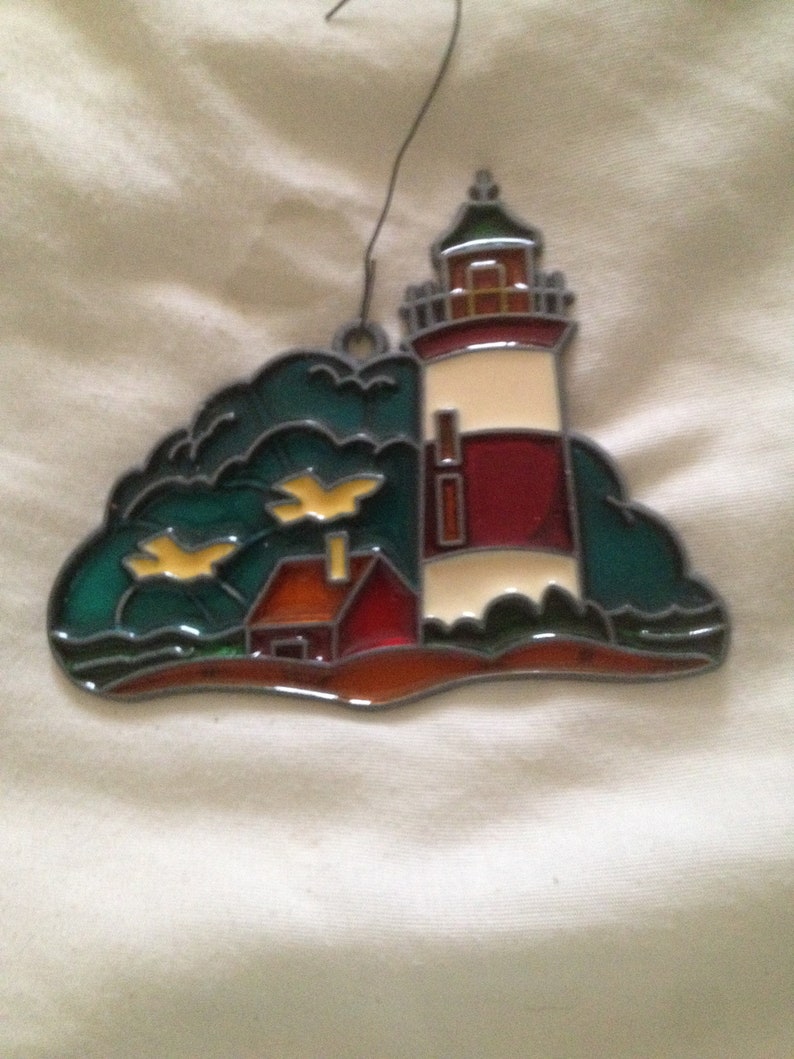 May include: A stained glass ornament depicting a red and white lighthouse with a small house in front of it. The lighthouse is on a small island with green trees and yellow birds flying overhead.