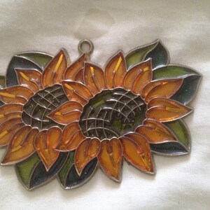 May include: Two stained glass sunflowers with orange and yellow petals and green leaves. The sunflowers are joined together at the center.