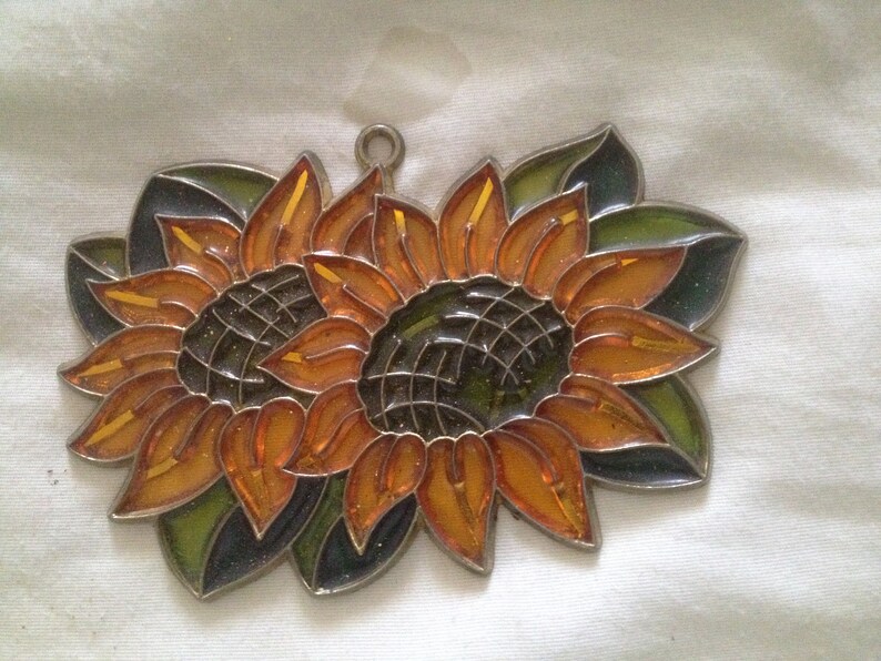 May include: Two stained glass sunflowers with orange and yellow petals and green leaves. The sunflowers are joined together at the center.
