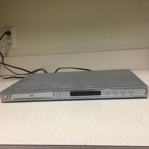 May include: A silver DVD player with a black display and buttons. The player has a slot for a DVD disc and a power button on the left side. The player is sitting on a white countertop.