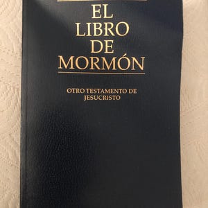 May include: A dark blue book with gold lettering. The text on the cover reads "EL LIBRO DE MORMON" and "OTRO TESTAMENTO DE JESUCRISTO". The book has a textured cover.