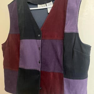 May include: A patchwork vest in shades of black, purple, and burgundy. The vest has a V-neck and a button-up front. The vest is hanging on a white hanger.