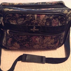 Puede incluir: Una bolsa de viaje negra con un diseño de tapicería floral y una correa de hombro negra ajustable. La bolsa tiene un cierre de cremallera y una placa de metal con el nombre "Atlantic".