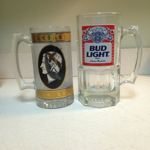 May include: Two glass beer mugs. The mug on the left has a gold trim and a picture of a man with a laurel wreath on his head. The mug on the right has a Bud Light logo and the words "Genuine" and "Genuine" on the sides.