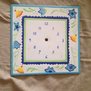 May include: Square, hand-painted clock face with a white background and blue numbers. The clock face is decorated with blue, yellow, and green floral designs and a blue border. The clock face has a hole in the center.