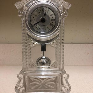 May include: A clear, quartz clock with Roman numerals on the face. The clock has a decorative, cut-glass design with a pendulum. The clock is rectangular with a peaked roof and ornate detailing.
