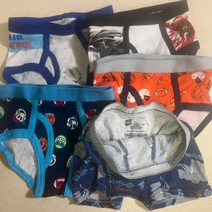 May include: A collection of children's underwear in various colors and patterns. Designs include sports equipment, cartoon characters, and the text "AIR CONTROL". The underwear has elastic waistbands and trim.