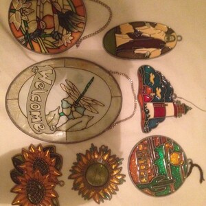 May include: Assortment of six stained glass suncatchers with various designs, including a dragonfly, sunflowers, a lighthouse, a cactus, and a welcome sign.