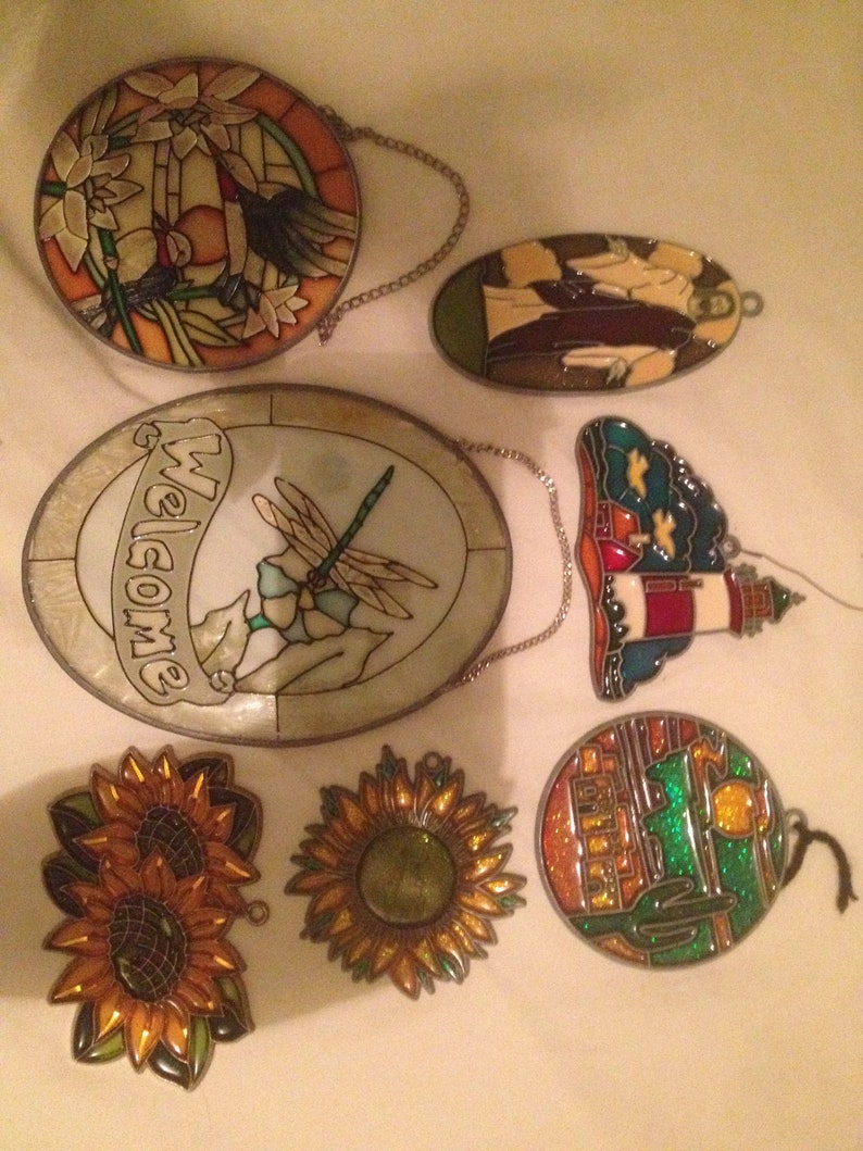 May include: Assortment of six stained glass suncatchers with various designs, including a dragonfly, sunflowers, a lighthouse, a cactus, and a welcome sign.