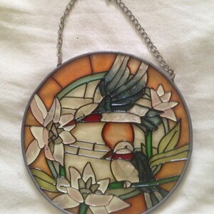 May include: A stained glass window hanging with a chain. The window features two birds, one flying and one perched on a branch, with flowers and leaves in shades of green, orange, and white.