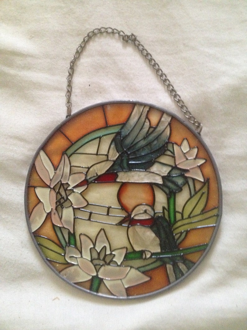 May include: A stained glass window hanging with a chain. The window features two birds, one flying and one perched on a branch, with flowers and leaves in shades of green, orange, and white.