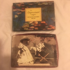 Może przedstawiać: Dwa pudełka na artykuły papiernicze w stylu vintage. Górne pudełko jest niebieskie z obrazem lilii wodnych i tekstem "Impressionist Stationery Box". Dolne pudełko jest czerwone z vintage'ową ilustracją dziewczyny w białej sukience trzymającej szczeniaka i pchającej wózek z dwoma szczeniakami w środku. Tekst na pudełku brzmi "Yestoryears".