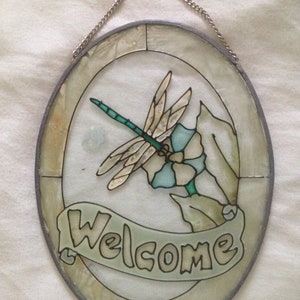 May include: A round stained glass window with a silver frame and a chain for hanging. The window features a dragonfly with blue and green wings, a blue flower, and green leaves. The words "Welcome" are written in a banner below the dragonfly.