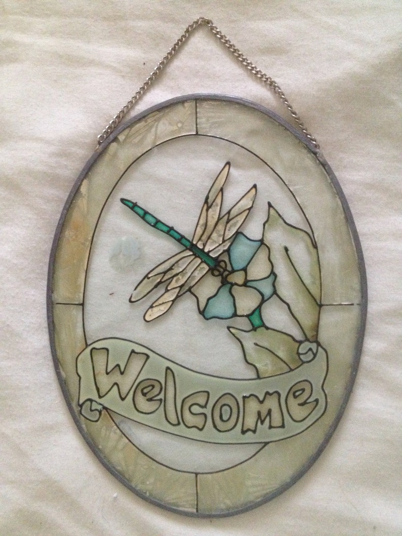 May include: A round stained glass window with a silver frame and a chain for hanging. The window features a dragonfly with blue and green wings, a blue flower, and green leaves. The words "Welcome" are written in a banner below the dragonfly.