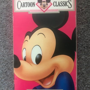 May include: A pink VHS tape with a cartoon image of Mickey Mouse. The tape is labeled "Walt Disney Cartoon Classics" and "Here's Mickey!"