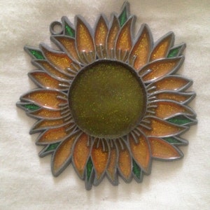 May include: A metal sunflower ornament with a glittery orange and green finish. The sunflower has a round center and is designed to hang.