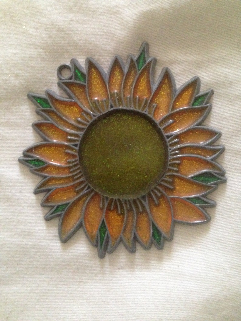 May include: A metal sunflower ornament with a glittery orange and green finish. The sunflower has a round center and is designed to hang.