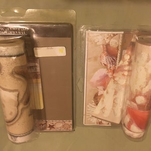 May include: Two rolls of beige and white wallpaper with a seashell design. The rolls are packaged in clear plastic and labeled "15 ft x 9.68 in (152.4 cm x 24.5 cm)".