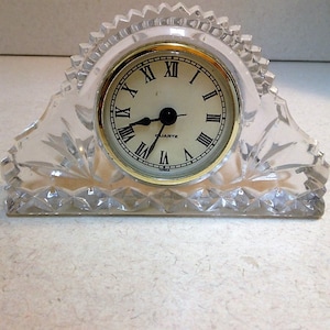 May include: A clear, crystal quartz clock with a gold-tone bezel and Roman numeral hour markers. The clock has a decorative, scalloped edge and a rectangular base. The clock face is cream-colored.