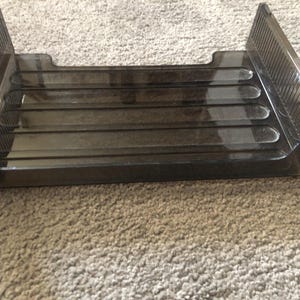 May include: A clear, rectangular desk organizer with a smoky gray tint. The organizer has a tray with four sections and two upright sides with a ribbed design. The item is designed to hold papers and other office supplies.