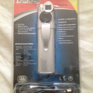 Puede incluir: Una cámara digital plateada con un puerto USB y un cable negro. La cámara está empaquetada en una carcasa de plástico transparente con un fondo azul y rojo. El empaque incluye el texto "DIGGERS" y "ArcSDf! es una marca registrada de Arcsoft Corporation".