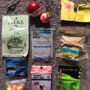 May include: A collection of fishing supplies, including floats, hooks, and bait. The image features various packaged items, such as wire leaders, trout bait rigs, and fluorescent fishing floats. Some items have text, including "FLUKE rig" and "YOU'RE".