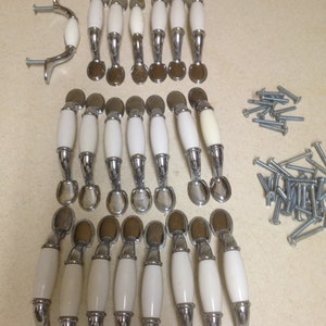Könnte beinhalten: Ein Set mit 20 weißen Keramik-Schrankknöpfen mit silbernen Akzenten. Die Knöpfe haben verschiedene Formen, darunter ovale und runde. Das Set enthält auch Schrauben für die Installation.