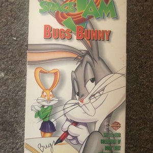 May include: VHS video cassette cover featuring Bugs Bunny from the movie Space Jam. The cover is white with a blue and red Space Jam logo and the text "Stars of Space Jam Bugs Bunny". The image shows Bugs Bunny drawing a heart with a red pen and a female bunny looking at him with hearts in her eyes.