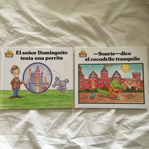 May include: Two colorful children's books with Spanish text. The book on the left has a picture of a man with a magnifying glass looking at a castle. The book on the right has a picture of a crocodile smiling and running.