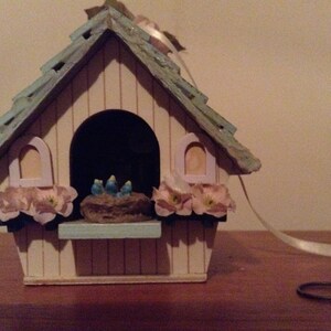 May include: A small wooden birdhouse painted in white and light blue with a pink and white flower trim. The birdhouse has a small opening for birds to enter and a small ledge for them to perch on. There are three blue birds inside the birdhouse.