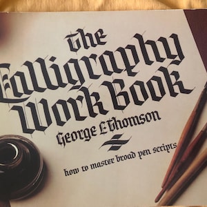 May include: The Calligraphy Work Book by George L. Thomson, featuring broad pen scripts. The cover shows a calligraphy inkwell, pens, and the book title in a gothic font. The book is on a cream-colored background.