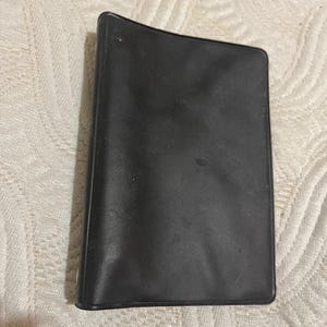 May include: A black, rectangular, leather-like wallet. The wallet is closed and has a slightly curved shape. The surface appears smooth with subtle variations in texture. The item is likely designed for carrying cards and cash.