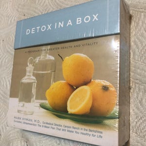 May include: A boxed set titled "DETOX IN A BOX" with the subtitle "A PROGRAM FOR GREATER HEALTH AND VITALITY." The cover features a still life of lemons on a green plate, a glass bottle, and a glass of water. The author's name and credentials are listed.