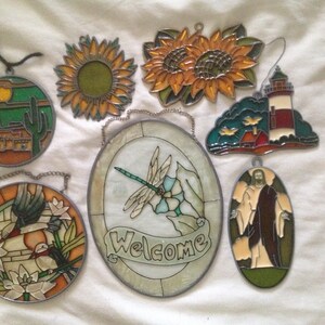 May include: Assortment of eight stained glass window hangings with various themes, including a sunset, a lighthouse, a dragonfly, a bird, a sunflower, and a religious figure.  The hangings are all made of colorful glass and have a metal frame.