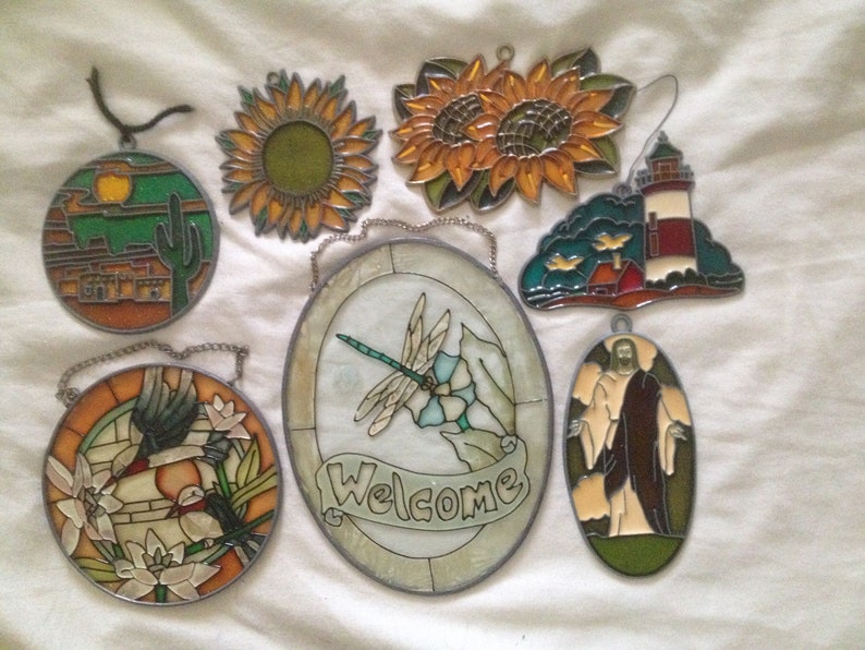 May include: Assortment of eight stained glass window hangings with various themes, including a sunset, a lighthouse, a dragonfly, a bird, a sunflower, and a religious figure.  The hangings are all made of colorful glass and have a metal frame.