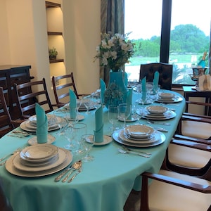 May include: A formal dining room table setting with a light blue tablecloth, white plates, silver cutlery, and glasses. The table is set for a large dinner party with multiple place settings. The room has a large window with a view of a green lawn and trees.