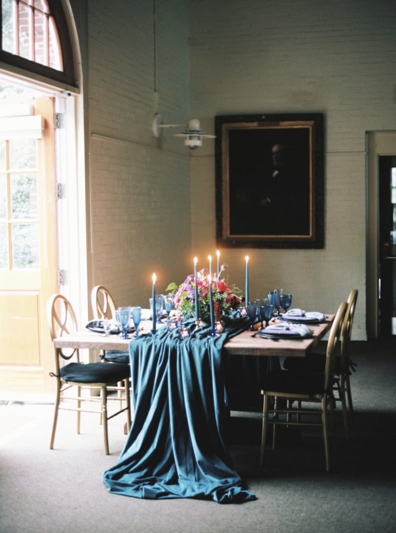 A teal blue velvet table runner drapes over a wooden table set for a dinner party. The table is decorated with a centerpiece of pink and purple flowers, blue candles, and blue glassware.