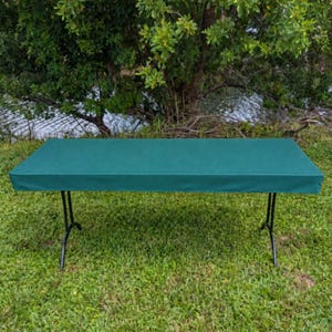 May include: A rectangular folding table covered with a solid dark green fitted tablecloth. The table features thin black metal legs that extend outward to support the flat surface, creating a clean and uniform appearance for the tabletop.