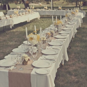 A long table set for a wedding reception outdoors. The table is covered in a white tablecloth with a burlap table runner down the center. There are white plates, silverware, and glasses set on the table. There are yellow flowers in a vase in the center of the table.