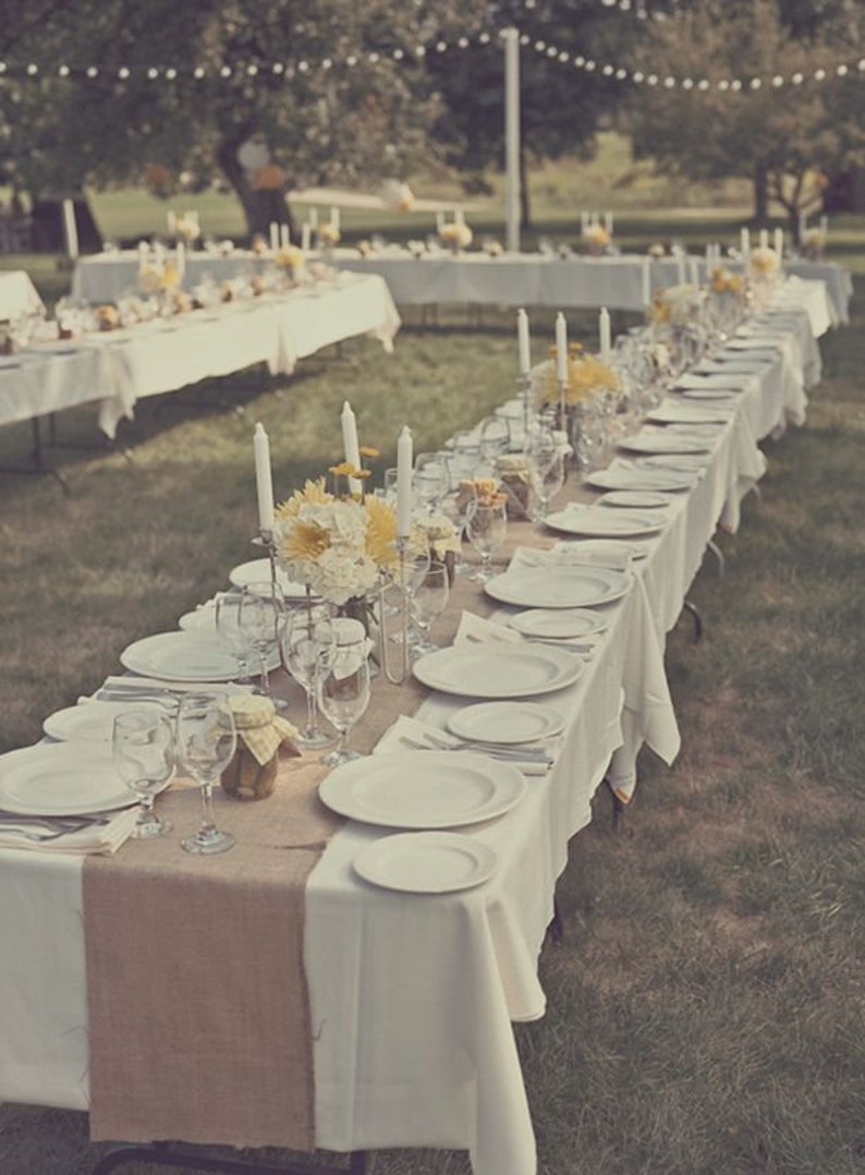 A long table set for a wedding reception outdoors. The table is covered in a white tablecloth with a burlap table runner down the center. There are white plates, silverware, and glasses set on the table. There are yellow flowers in a vase in the center of the table.