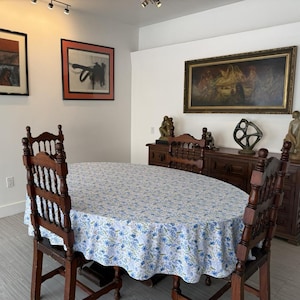May include: A dining room set with a round table covered in a blue and white floral tablecloth. Two wooden chairs are visible, along with artwork and a wooden cabinet in the background. The room has white walls and a light-colored floor.