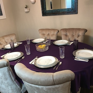 May include: A round dining table with a purple tablecloth and six chairs. The table is set with white plates, silver chargers, silverware, and glasses. There is a bowl of lemons in the center of the table.