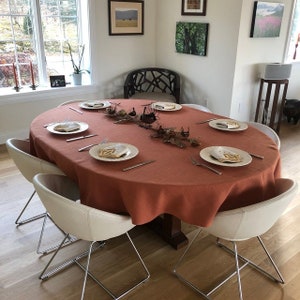May include: A round, orange linen tablecloth is set on a wooden table with six white chairs. The table is set with six place settings, each with a white plate, a gold napkin, and a fork and knife. There is a centerpiece of gold branches and leaves in the center of the table.