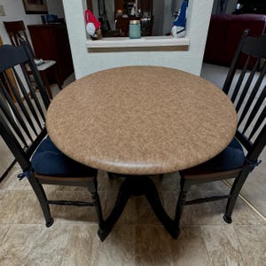 May include: A round dining table with a textured tan surface and a black pedestal base, accompanied by two black wooden chairs featuring vertical slats and dark blue seat cushions tied with fabric ribbons at the back of each chair frame.