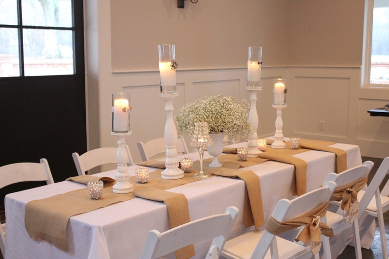 A  table with a white tablecloth and burlap runners. There are white candlesticks with white candles, a vase with white flowers, and small glass candle holders with tea lights. There are also white chairs around the table.