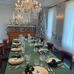 A formal dining room table setting with a large green velvet tablecloth, gold chargers, white dinner plates with a green Christmas tree design, green velvet napkins, and silver flatware.  The table linen is a wrinkle free tablecloth. There are clear glasses and water glasses on the table. The table is set for a holiday dinner with a silver chandelier hanging above and a Christmas tree in the background.