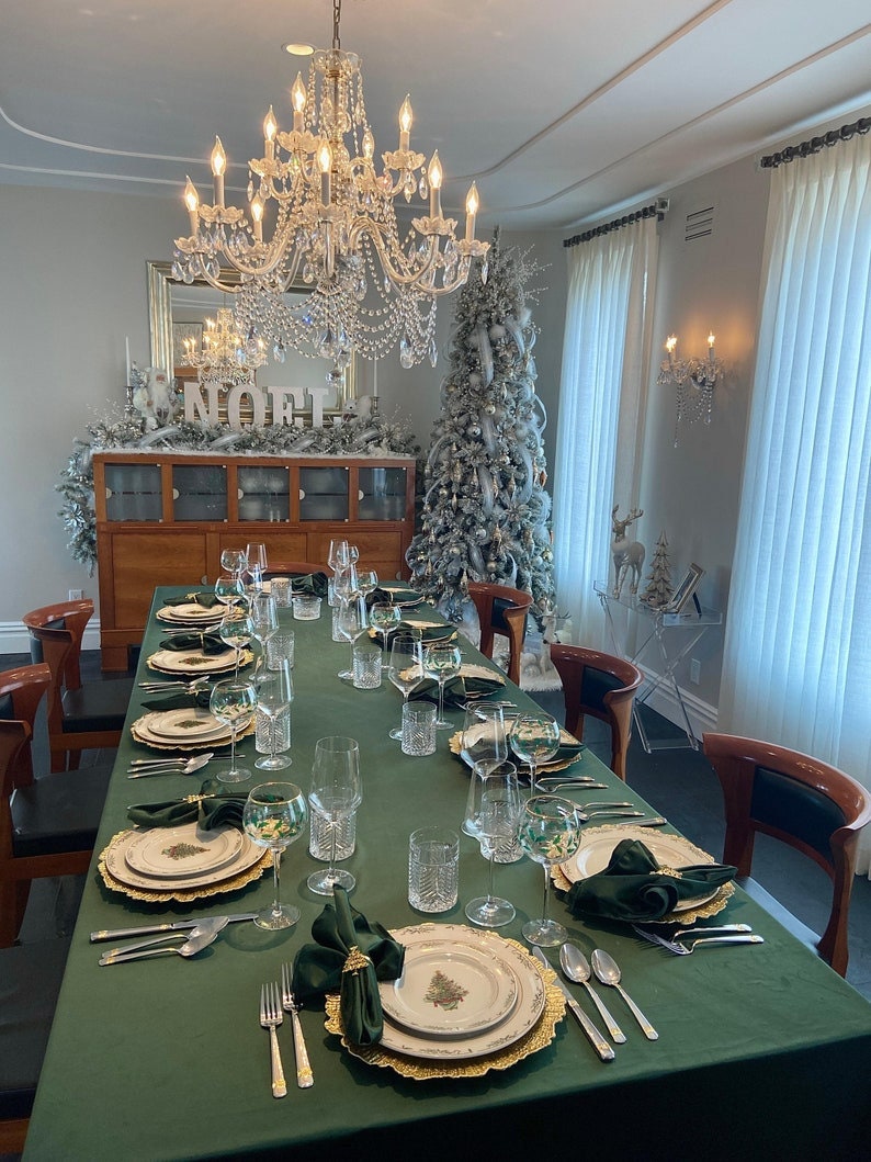 A formal dining room table setting with a large green velvet tablecloth, gold chargers, white dinner plates with a green Christmas tree design, green velvet napkins, and silver flatware.  The table linen is a wrinkle free tablecloth. There are clear glasses and water glasses on the table. The table is set for a holiday dinner with a silver chandelier hanging above and a Christmas tree in the background.